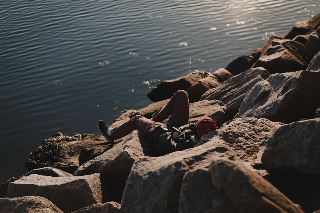 person in black pants sitting on gray rock near body of water during daytime