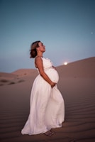 A pregnant woman in a flowing white dress stands on sand dunes under a twilight sky. The moon is visible in the background, adding to the serene atmosphere.