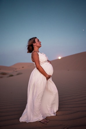 A pregnant woman in a flowing white dress stands on sand dunes under a twilight sky. The moon is visible in the background, adding to the serene atmosphere.