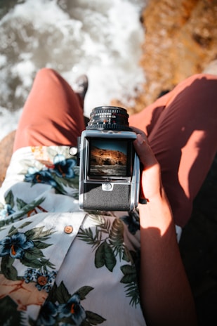 A person sitting by the water holds a vintage camera focused on capturing a scenic landscape. The person wears a tropical shirt with floral patterns and reddish-brown pants. The background shows waves splashing against rocks.