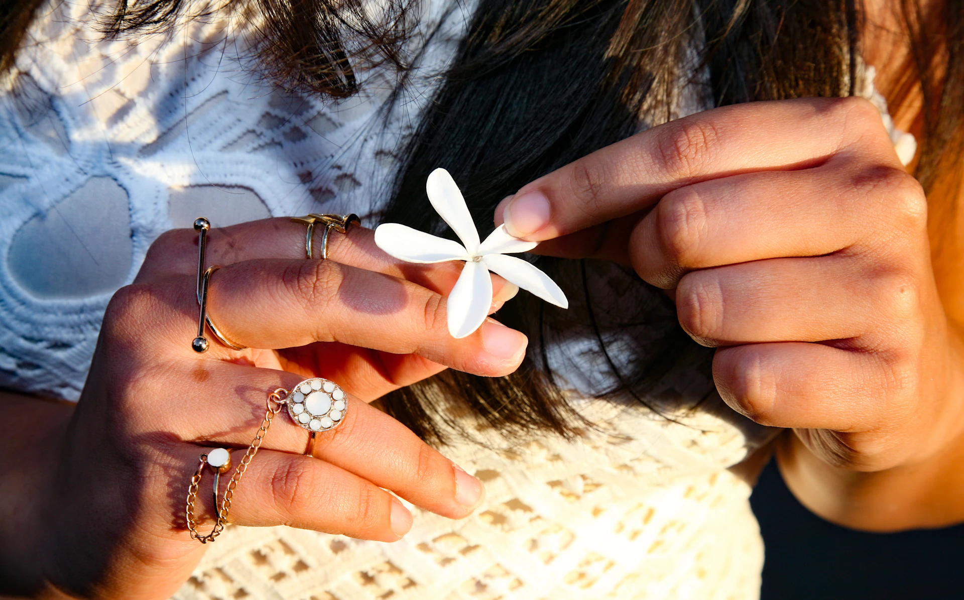 A hand adorned with several rings featuring colorful gemstones, gently holding a blooming flower in soft focus.