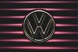A metallic Volkswagen logo is prominently displayed on a background of horizontal maroon lines. The lighting creates a dramatic and focused effect on the emblem, giving it a striking presence.