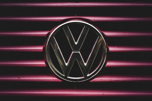 A metallic Volkswagen logo is prominently displayed on a background of horizontal maroon lines. The lighting creates a dramatic and focused effect on the emblem, giving it a striking presence.