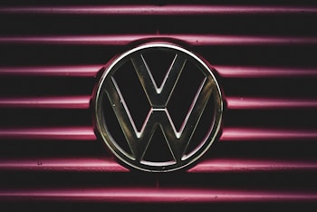 A metallic Volkswagen logo is prominently displayed on a background of horizontal maroon lines. The lighting creates a dramatic and focused effect on the emblem, giving it a striking presence.