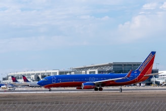 A Southwest Airlines plane is taxiing on the tarmac of an airport. The aircraft is painted in the airline's signature blue, red, and orange colors. In the background, several Delta Airlines planes are parked near a modern airport terminal building. The sky is partly cloudy.