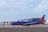 A Southwest Airlines plane is taxiing on the tarmac of an airport. The aircraft is painted in the airline's signature blue, red, and orange colors. In the background, several Delta Airlines planes are parked near a modern airport terminal building. The sky is partly cloudy.