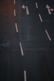 A dark asphalt road with several white lane markings and directional arrows. There is an arrow pointing to the left and another pointing straight. The surface appears slightly wet or reflective, suggesting recent rain or moisture.