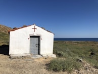 A small, white chapel with a red-tiled roof is situated on a grassy hillside overlooking the sea. The building has a simple rectangular structure, adorned with a red cross above the door. The landscape around the chapel is natural and unspoiled, with green vegetation and rocks leading to the serene blue expanse of the ocean in the background under a clear blue sky.