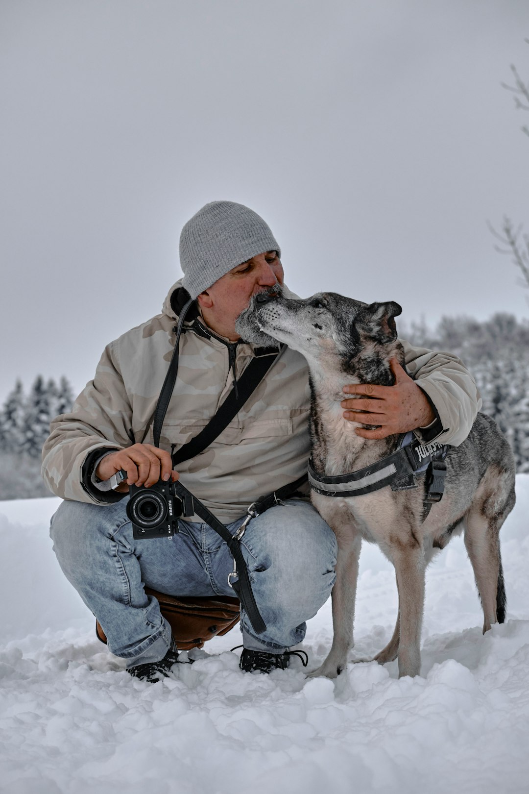 man in brown jacket and blue denim jeans sitting on snow covered ground with dog during