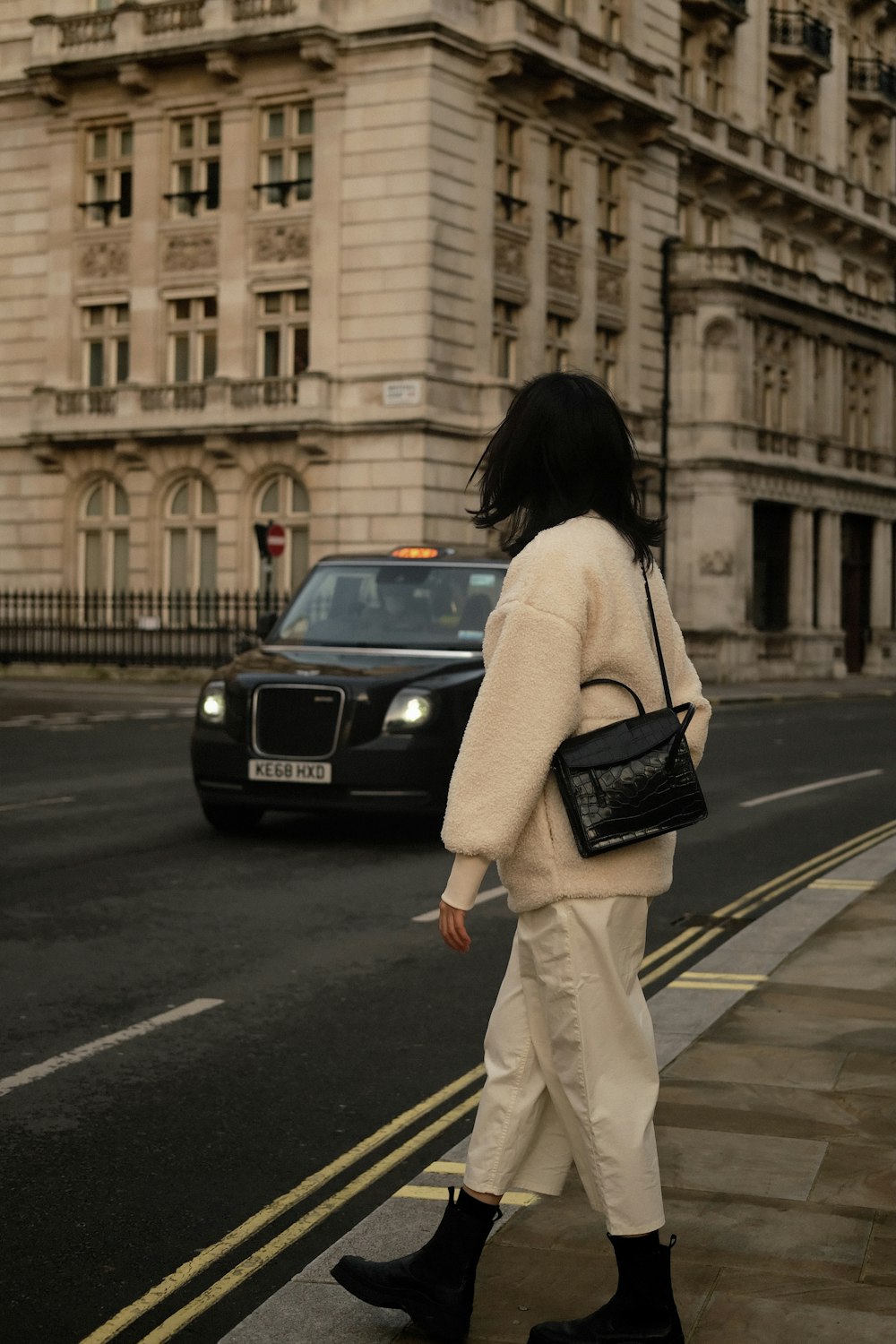 femme en chemise blanche à manches longues et pantalon blanc tenant un sac à main en cuir noir marchant sur un piéton