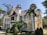 A grand, ornate building with tall, pointed red roofs and intricate architectural details, reminiscent of a castle or historic mansion. Surrounded by lush greenery, including palm trees and manicured bushes, indicating a well-maintained garden. The structure features white walls with decorative columns and multiple balconies.