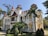 A grand, ornate building with tall, pointed red roofs and intricate architectural details, reminiscent of a castle or historic mansion. Surrounded by lush greenery, including palm trees and manicured bushes, indicating a well-maintained garden. The structure features white walls with decorative columns and multiple balconies.