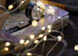 A miniature white car is placed on a small model road, which is part of a book or journal setup with an open notebook. Nearby is a roll of black tape designed to resemble a road. Warm, glowing fairy lights are scattered around, creating a cozy and whimsical ambiance.