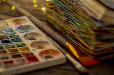 A well-used watercolor palette with various paint colors and brushes is positioned next to a stack of colorful paper. The colors are rich and vibrant, indicating frequent use. The background is softly blurred with small warm lights providing a cozy ambiance.