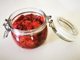 A glass jar filled with a chunky red preserve, possibly strawberry jam, sits on a light surface. The jar has a metal hinge and a clear lid that is slightly open, revealing the vibrant texture and color of the contents.