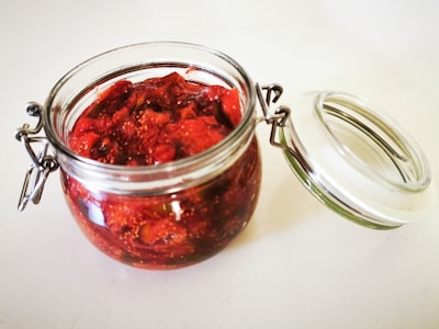 A glass jar filled with a chunky red preserve, possibly strawberry jam, sits on a light surface. The jar has a metal hinge and a clear lid that is slightly open, revealing the vibrant texture and color of the contents.