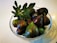 A clear glass bowl filled with ripe, dark purple figs, some with a hint of green. A fresh fig leaf is placed on top of the figs, adding a touch of greenery to the arrangement. The white background enhances the vibrant colors of the fruit and the leaf.