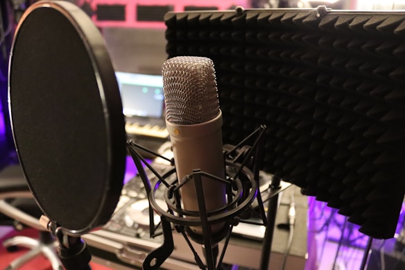 A professional recording setup featuring a large microphone with a pop filter and a background of acoustic foam panels. The setting includes dim lighting with purple hues and a glimpse of studio equipment.