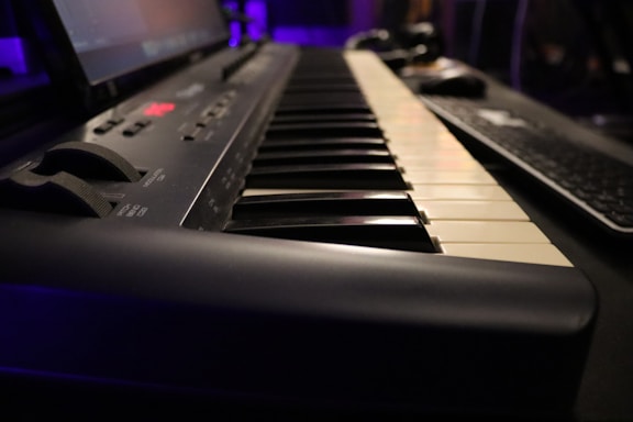 A close-up perspective of a digital keyboard with black and white keys visible. The focus is on the keys, while the background contains a blurred computer keyboard and a purple-lit setting, suggesting a desktop or studio environment.