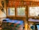 A cozy, rustic room with wooden walls and ceiling, featuring a single bed covered with a colorful quilt. The room is adorned with a series of vintage pennants hanging from the ceiling, adding a nostalgic feel. Large windows let in ample natural light, revealing a view of trees outside. A wooden desk with a book and a small plant is positioned under one of the windows. Traditional snowshoes and a wooden paddle are also visible, emphasizing a classic, outdoorsy theme.