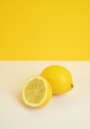 Two lemons are placed on a simple background, with one lemon sliced in half to reveal the juicy interior. The vibrant yellow of the lemons complements the bold yellow background.