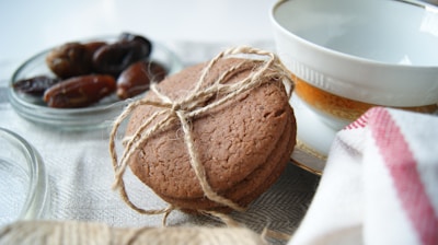 A stack of brown cookies tied with string is placed next to a white teacup with a gold rim. There is a small glass bowl containing dates in the background, and a white cloth with a red stripe is partially visible. The scene is set on a light-textured fabric surface.