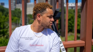 A person with curly hair is wearing a white t-shirt that has the words 'Love Disorder' printed in red. The background features a blurred view of greenery and buildings behind a brown metal structure.