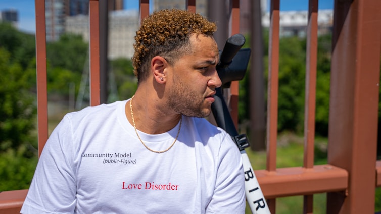 A person with curly hair is wearing a white t-shirt that has the words 'Love Disorder' printed in red. The background features a blurred view of greenery and buildings behind a brown metal structure.