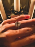 A hand is adorned with an elegant teardrop-shaped diamond ring. The background features candles and decorative greenery, suggesting a festive or intimate setting. The lighting creates warm hues on the skin and highlights the sparkle of the diamond.