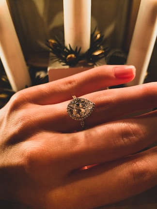 A hand is adorned with an elegant teardrop-shaped diamond ring. The background features candles and decorative greenery, suggesting a festive or intimate setting. The lighting creates warm hues on the skin and highlights the sparkle of the diamond.