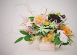 A detailed and ornate floral arrangement displayed in a white rectangular container. The arrangement includes a variety of flowers and green foliage, with colors that range from soft pastels to deep rich hues. The flowers include pink, yellow, and dark red blooms along with succulents and white cotton-like elements, all accompanied by green leaves and delicate twigs.