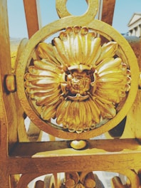 Ornate metalwork featuring a floral design prominently displayed in golden hues. The flower motif is set within a circular framework, reflecting sunlight that highlights its intricate details.