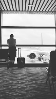 A person stands at an airport window looking out at an airplane on the tarmac. The individual is accompanied by a suitcase and the airport terminal has a patterned carpet floor. Overhead lights and ceiling beams add to the structured environment.