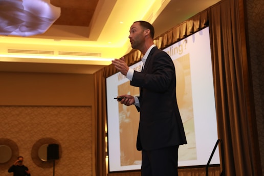 A person in formal attire is giving a presentation in a conference room. The person is holding a device, possibly a remote, and gesturing while speaking. A large projection screen is visible in the background, accompanied by elegant lighting and decor.