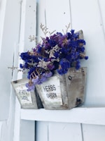 A rustic wooden herb planter is mounted on a light-painted wooden background. The planter holds dried purple flowers and brown twigs, creating a charming, natural display. The words 'HERBS GARDEN' are printed on the planter's surface.