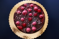 A round chocolate cake decorated with vibrant red cherries, pink frosting florets, and small silver sugar pearls. The cake rests on a golden cake board, providing an elegant contrast against the dark brown cake and decorations.
