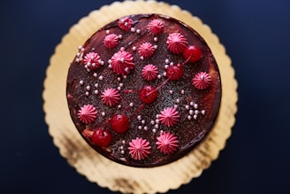 A round chocolate cake decorated with vibrant red cherries, pink frosting florets, and small silver sugar pearls. The cake rests on a golden cake board, providing an elegant contrast against the dark brown cake and decorations.