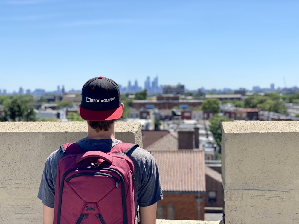 Mann im blauen Rundhals-T-Shirt mit rot-schwarzem Rucksack auf braunem Beton stehend