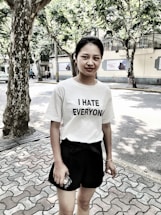 A person stands outdoors on a paved walkway next to a tree. They are wearing a white t-shirt with the text 'I HATE EVERYONE' and black shorts, holding a mobile phone. The background shows trees providing shade and a building across the street.