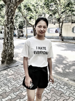 A person stands outdoors on a paved walkway next to a tree. They are wearing a white t-shirt with the text 'I HATE EVERYONE' and black shorts, holding a mobile phone. The background shows trees providing shade and a building across the street.