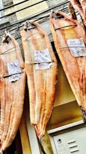 Large fish are hung side by side, drying with labels attached to each. The fish have a pinkish hue and are displayed against an urban backdrop with buildings.