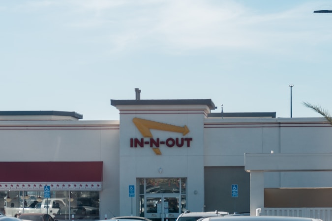 A fast-food restaurant with a prominent red and yellow sign featuring an arrow. The building has a beige facade and a red awning above the windows. Several cars are parked in front of the entrance, and there are handicap parking signs visible.