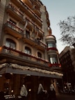 A grand hotel building with intricate balconies and festive decorations, including lights and garlands. The facade is adorned with red flowers and various flags hanging from the balconies. The entrance is elegantly decorated with illuminated Christmas trees, creating a warm, inviting atmosphere. There are trees lining the street beside the hotel.