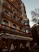 A grand hotel building with intricate balconies and festive decorations, including lights and garlands. The facade is adorned with red flowers and various flags hanging from the balconies. The entrance is elegantly decorated with illuminated Christmas trees, creating a warm, inviting atmosphere. There are trees lining the street beside the hotel.