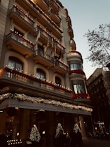 A grand hotel building with intricate balconies and festive decorations, including lights and garlands. The facade is adorned with red flowers and various flags hanging from the balconies. The entrance is elegantly decorated with illuminated Christmas trees, creating a warm, inviting atmosphere. There are trees lining the street beside the hotel.