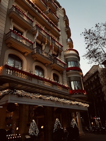 A grand hotel building with intricate balconies and festive decorations, including lights and garlands. The facade is adorned with red flowers and various flags hanging from the balconies. The entrance is elegantly decorated with illuminated Christmas trees, creating a warm, inviting atmosphere. There are trees lining the street beside the hotel.