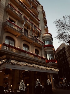 A grand hotel building with intricate balconies and festive decorations, including lights and garlands. The facade is adorned with red flowers and various flags hanging from the balconies. The entrance is elegantly decorated with illuminated Christmas trees, creating a warm, inviting atmosphere. There are trees lining the street beside the hotel.