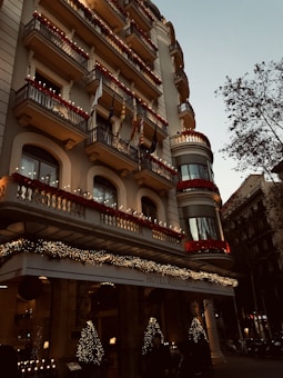 A grand hotel building with intricate balconies and festive decorations, including lights and garlands. The facade is adorned with red flowers and various flags hanging from the balconies. The entrance is elegantly decorated with illuminated Christmas trees, creating a warm, inviting atmosphere. There are trees lining the street beside the hotel.