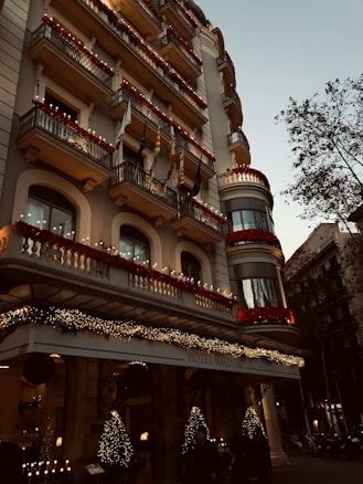 A grand hotel building with intricate balconies and festive decorations, including lights and garlands. The facade is adorned with red flowers and various flags hanging from the balconies. The entrance is elegantly decorated with illuminated Christmas trees, creating a warm, inviting atmosphere. There are trees lining the street beside the hotel.
