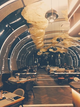 The image captures a modern and stylish restaurant interior with curved architectural lighting overhead. The setting features wooden tables and chairs, with ambient lighting creating a warm atmosphere. Unique yellow fabric sculptures hang from the ceiling, contributing to the contemporary aesthetic.